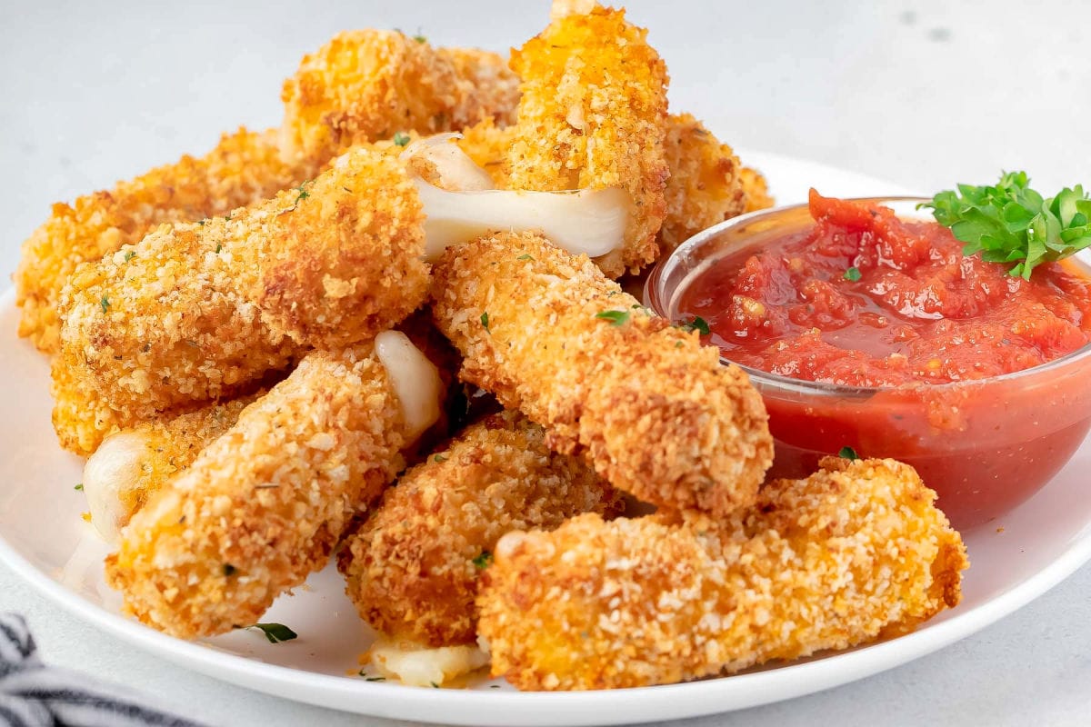 lots of mozzarella sticks cooked int he air fryer sitting on white plate with small bowl of marinara sauce.