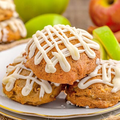 apple cookies sitting on white plate with gold rim with maple icing drizzle don top. fresh apples and apple slices in background.