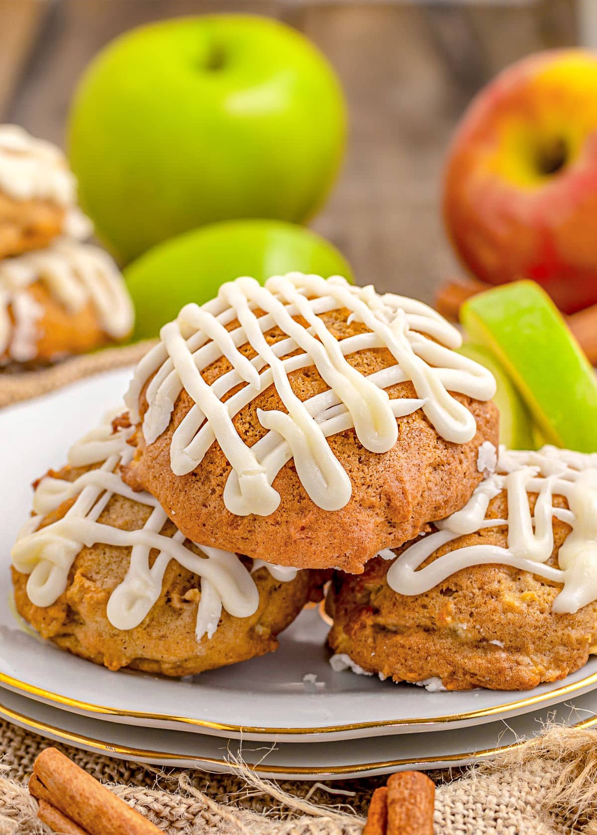 three apple cookies sitting on white plate with gold rim with maple icing drizzle don top. fresh apples and apple slices in background.