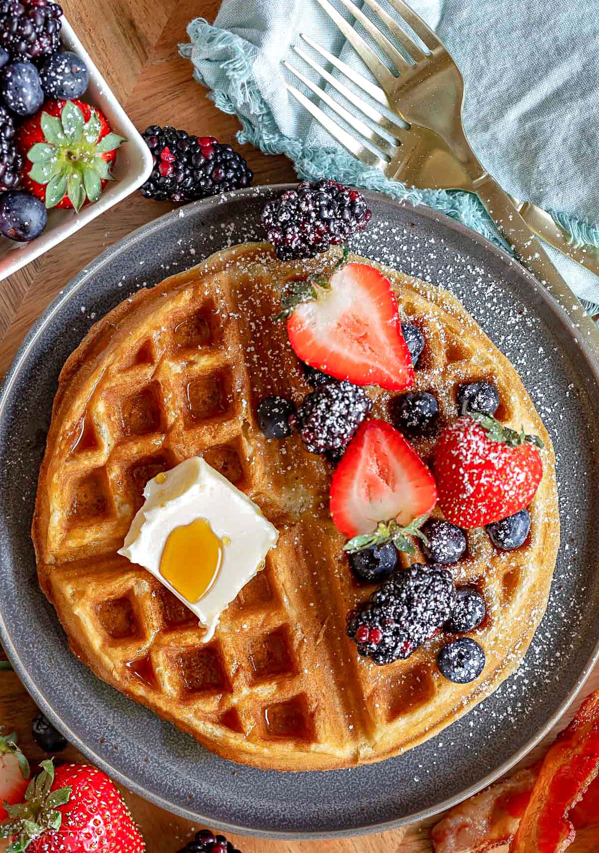 top down view of gray plate with one waffle. Half of the waffle topped with butter and syrup and half topped with berries and powdered sugar.