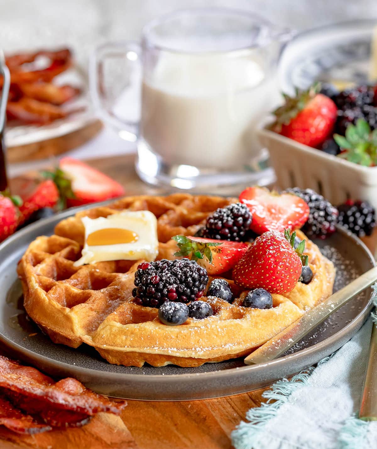 one waffle topped with berries and powdered sugar as well as some butter and syrup. milk and bacon in background.