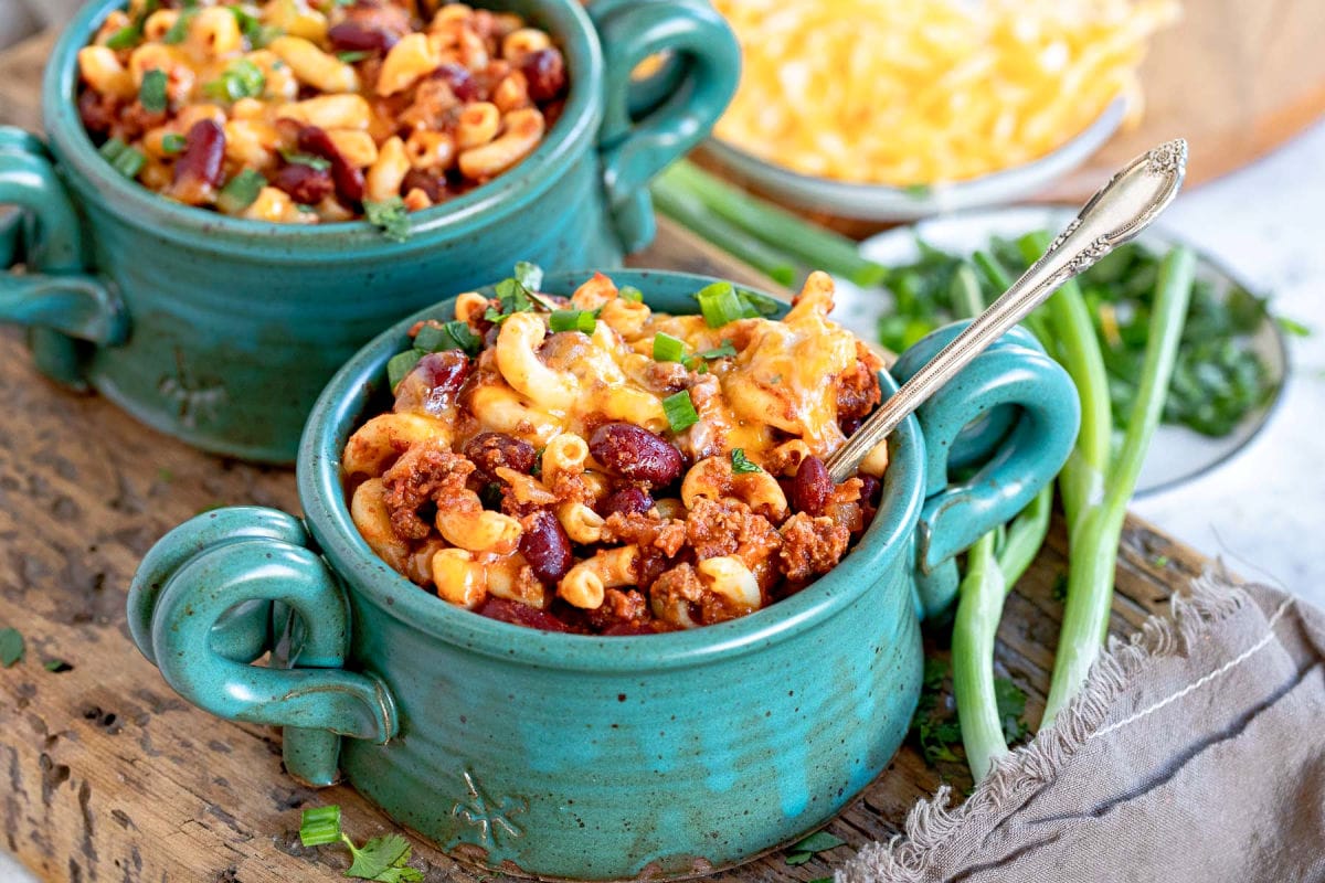 two bowls of chili mac topped with colby jack cheese and green onions. sitting on a thick wood board.