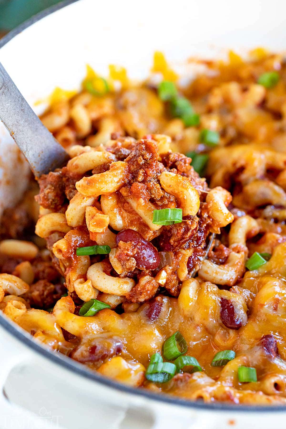 ladle of chili mac recipe in a white dutch oven topped with green onions.