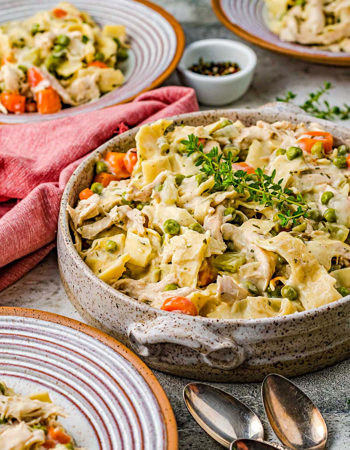 large pottery bowl filled with chicken noodles recipe topped with fresh thyme. Small dinner plates also have servings on them.