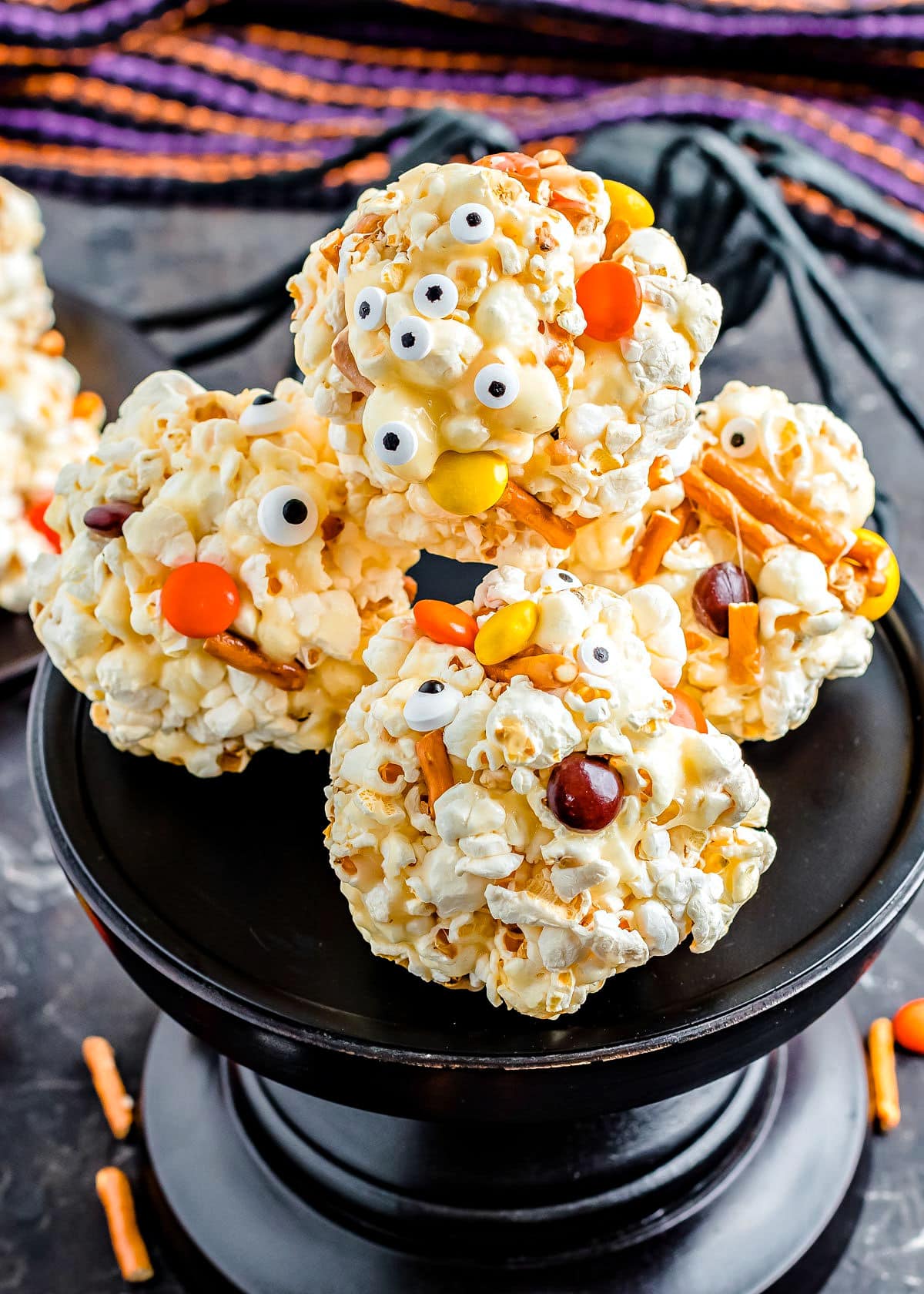 popcorn balls decorated for halloween with pretzels and candies sitting on black cake stand.