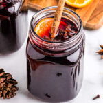 glass jar filled with spiced wine and topped with a cinnamon stick, orange slice, and star anise. sliced oranges in background.