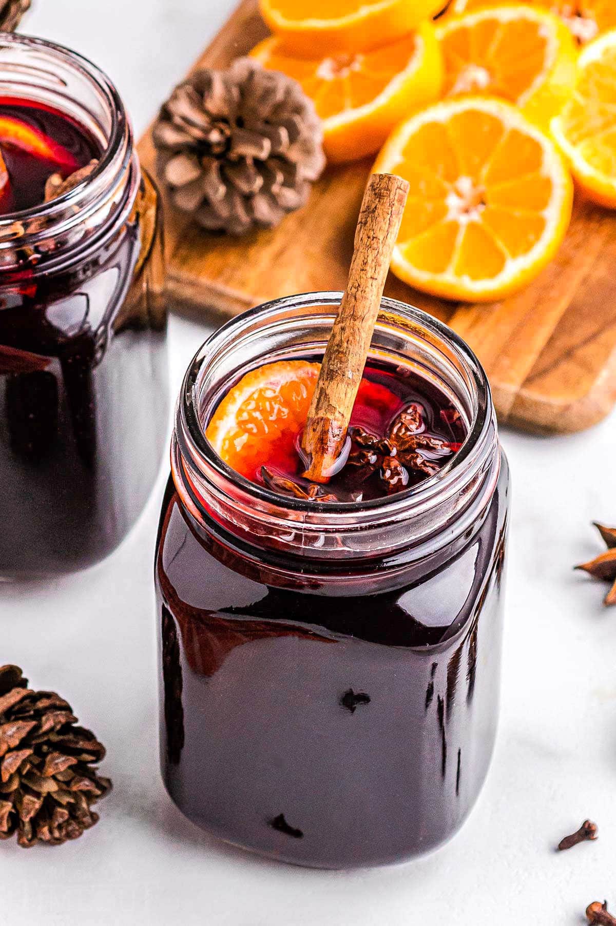 glass jar filled with spiced wine and topped with a cinnamon stick, orange slice, and star anise. sliced oranges in background.