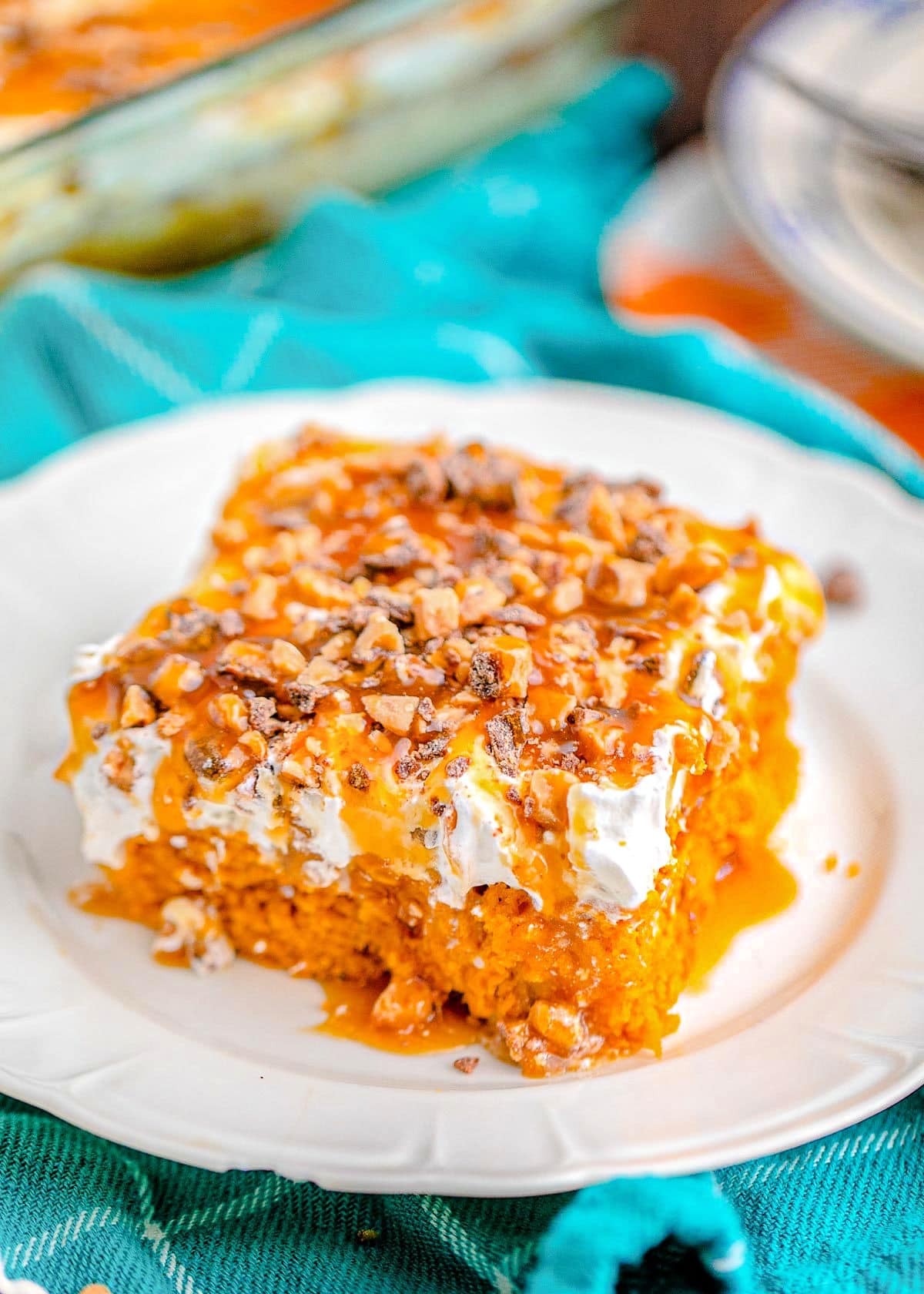 pumpkin cake sitting on white plate with bright blue napkin in the background. topped with cool whip and heath toffee bits.