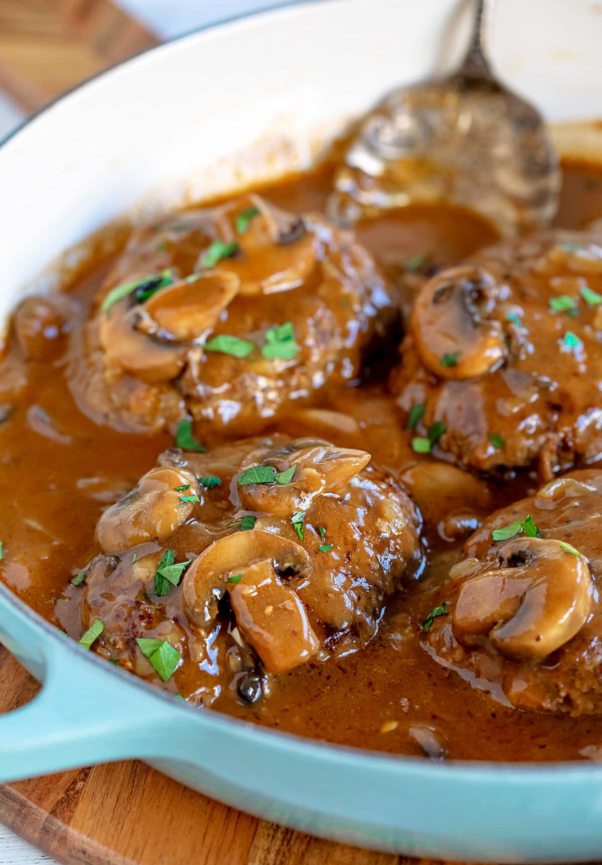 four salisbury steaks in large skillet with mushroom gravy and fresh parsley sprinkled over the top of them and antique spoon in background.