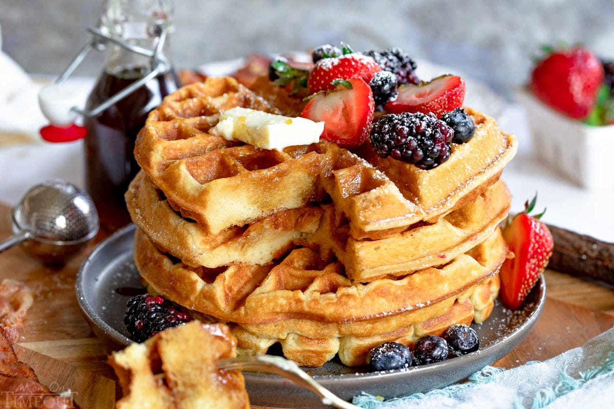 four waffles stacked on gray plate and topped with butter and berries. fork with bite on it sitting near plate.