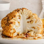 whole cauliflower head that has been seasoned and roasted in oven sitting on white plate with about one quarter of the head removed.