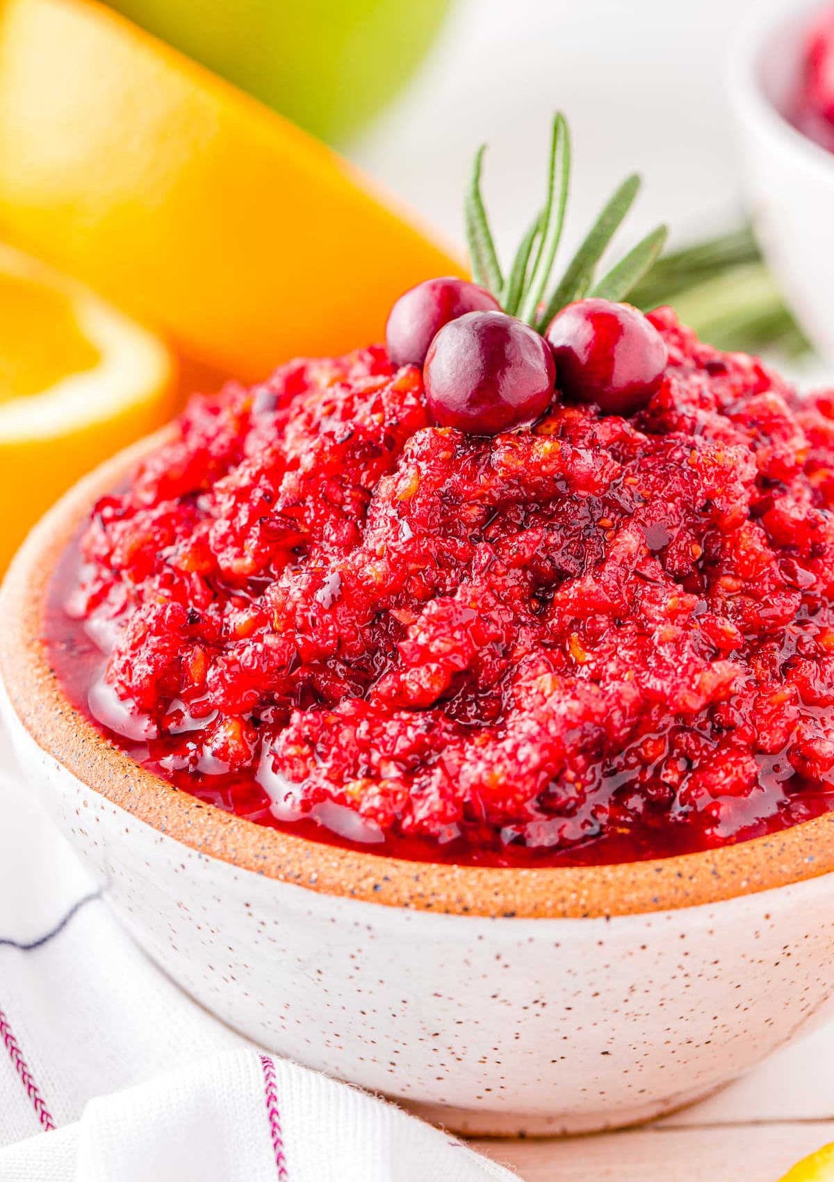 bowl of cranberry relish made with oranges and apples in a small white bowl topped with rosemary and fresh cranberries.