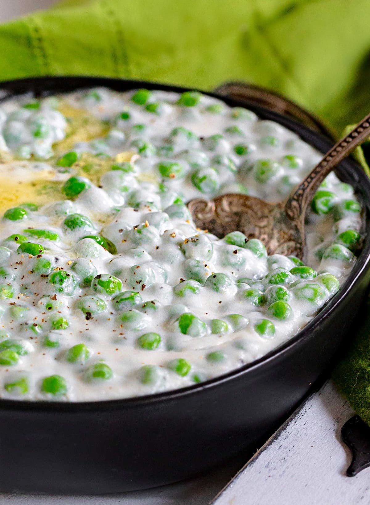 creamed peas in shallow black bowl with melted butter on top and antique serving spoon in bowl. green napkin in background.