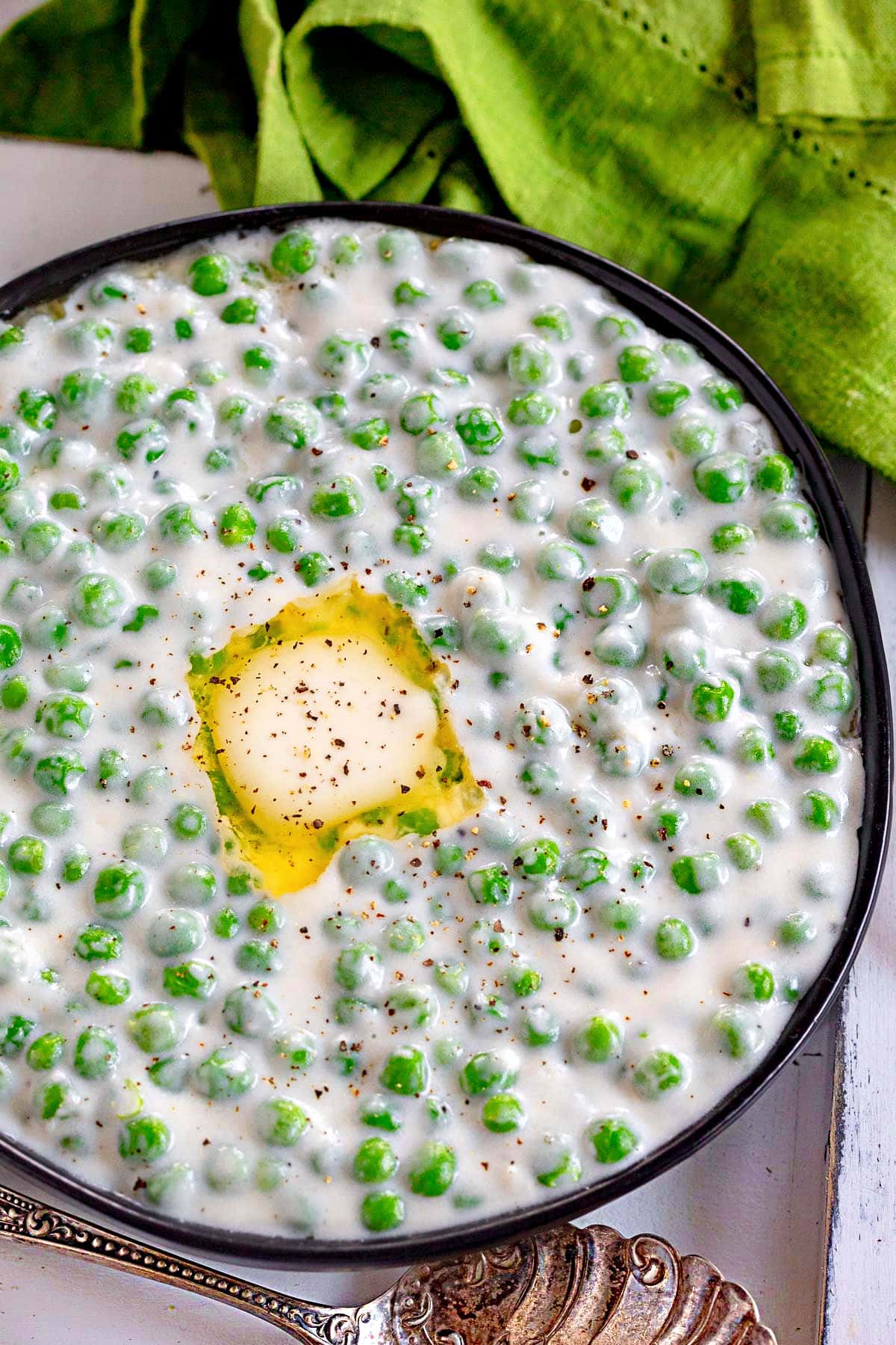 top down look at creamed peas in black bowl with melted butter and freshly cracked black pepper on top.