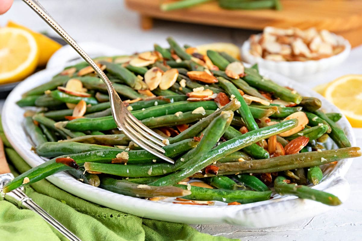 fork full of green beans almondine topped with toasted almonds on a shallow white serving dish with lemon slices scattered to the side sitting next to a green cloth napkin.