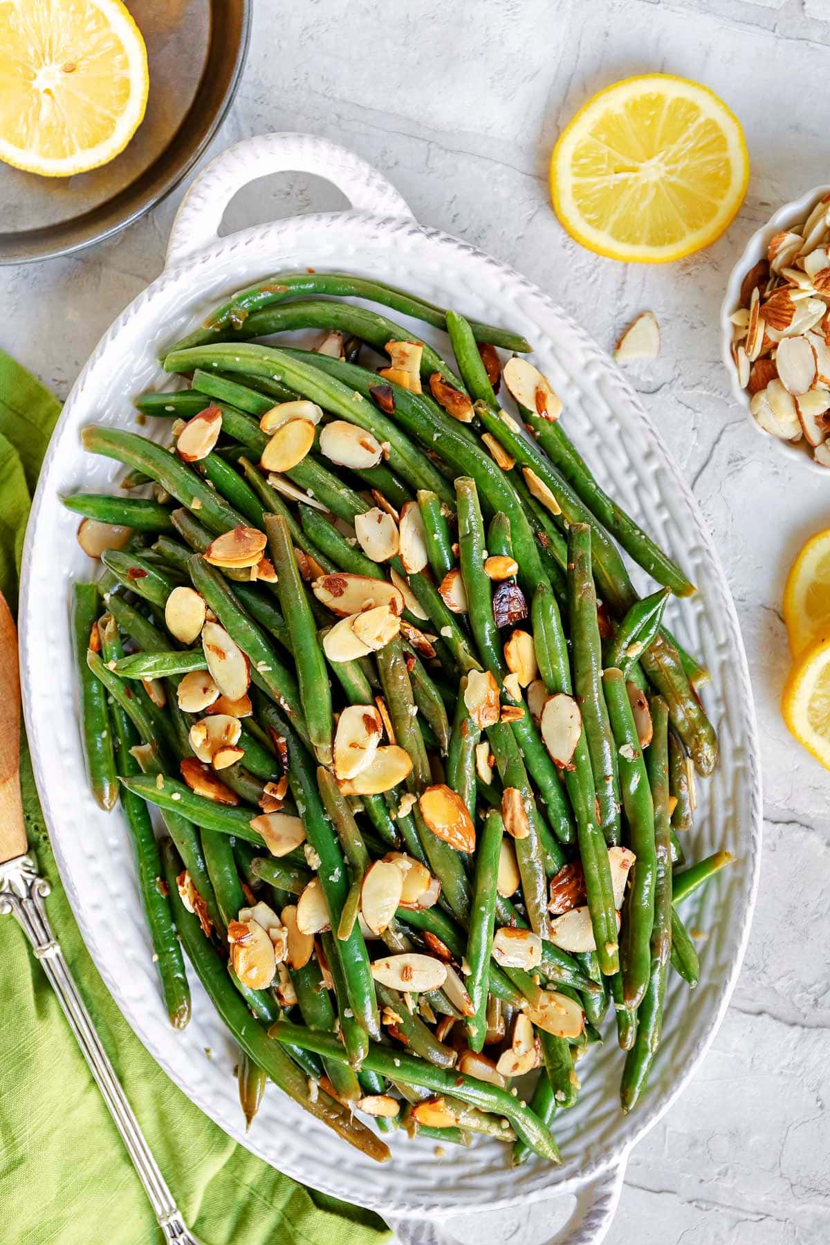 top down look at green beans almondine topped with toasted almonds on a shallow white serving dish with lemon slices scattered to the side.