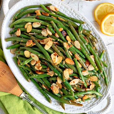 top down look at green beans almondine topped with toasted almonds on a shallow white serving dish with lemon slices scattered to the side.