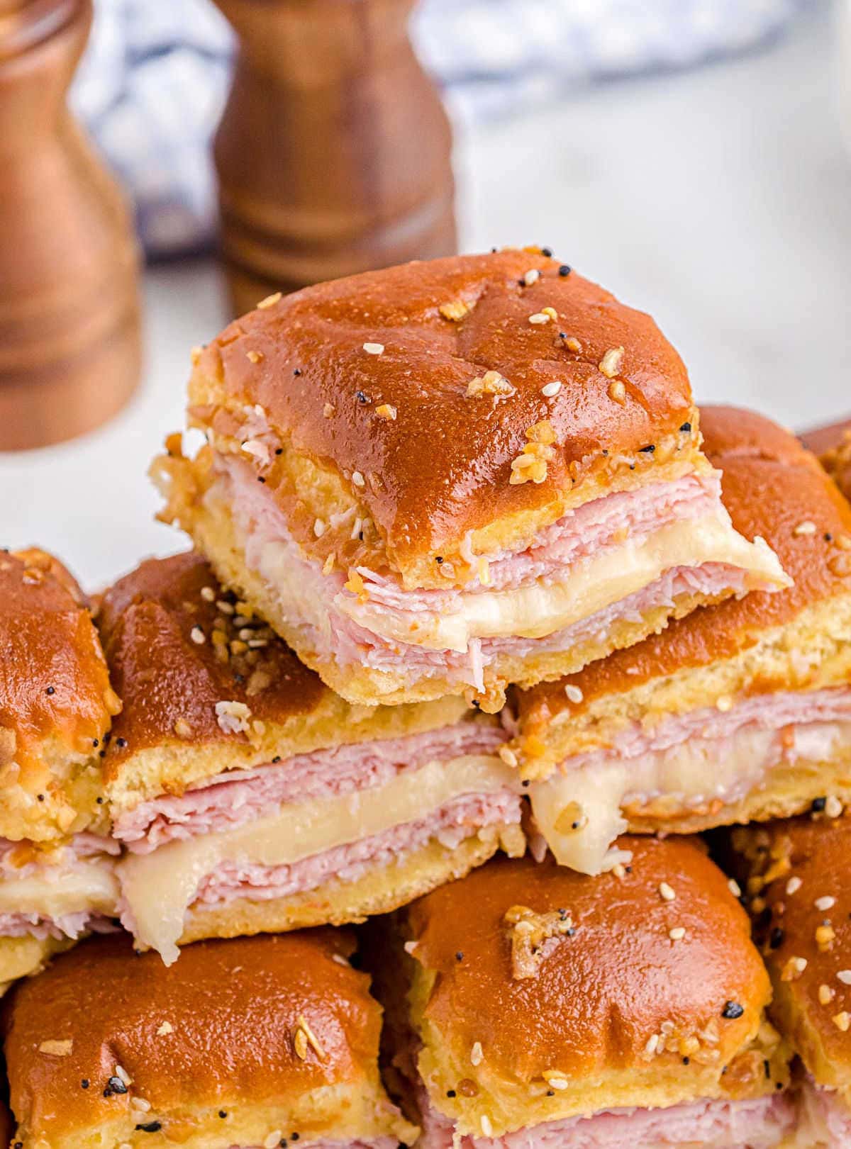 tall stack of hot ham and cheese sliders on serving platter ready to be enjoyed. wood salt and pepper shakers in background next to white and blue checked towel.