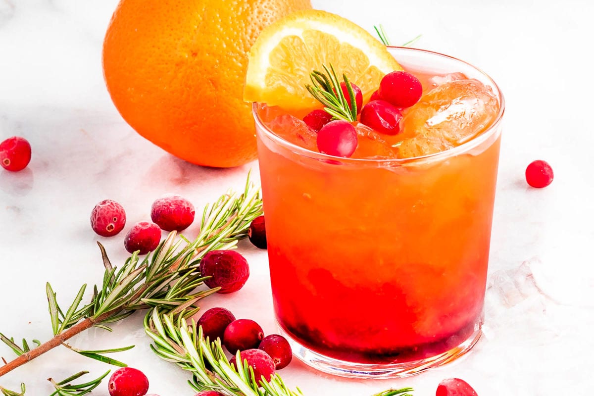 pretty crystal glass filled with christmas punch and garnished with rosemary, fresh cranberries and orange slice. more rosemary, cranberries, and an orange scattered to the side.