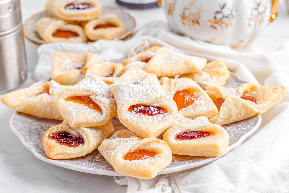 large china plate stacked high with kolachy cookies and dusted with powdered sugar. cookies have jam centers and are ready to be enjoyed.