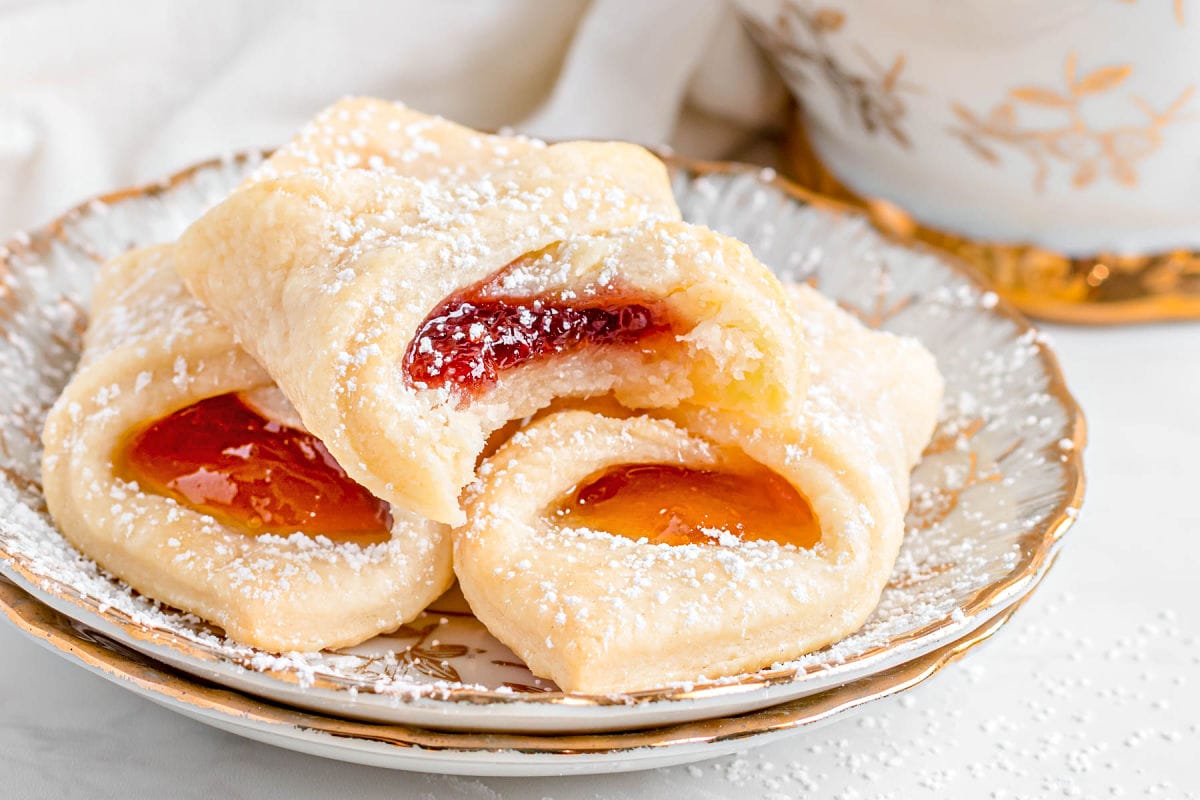 three kolaczki cookies sitting on a small plate with gold rim. the top cookie has a bite taken out of it.