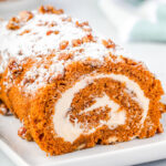 whole pumpkin cake roll on a white rectangle plate with powdered sugar and candied pecans on top of the cake roll.