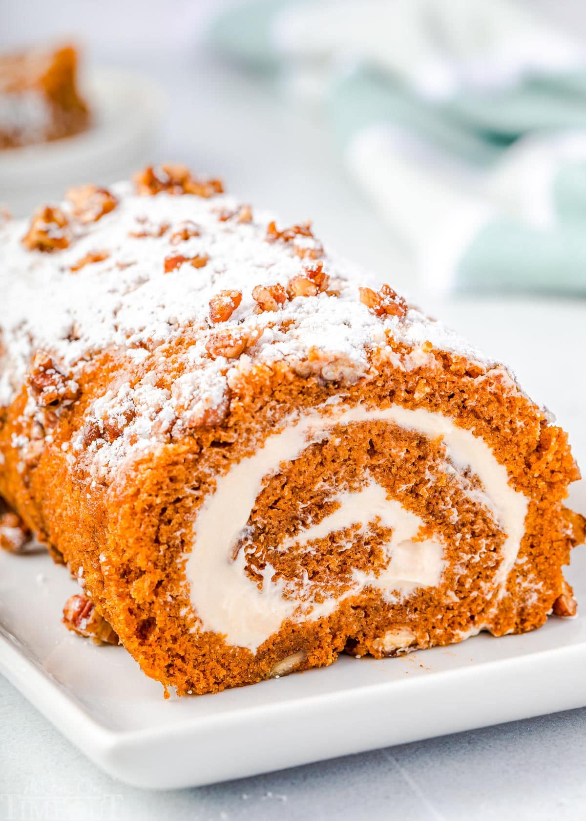 whole pumpkin cake roll on a white rectangle plate with powdered sugar and candied pecans on top of the cake roll.