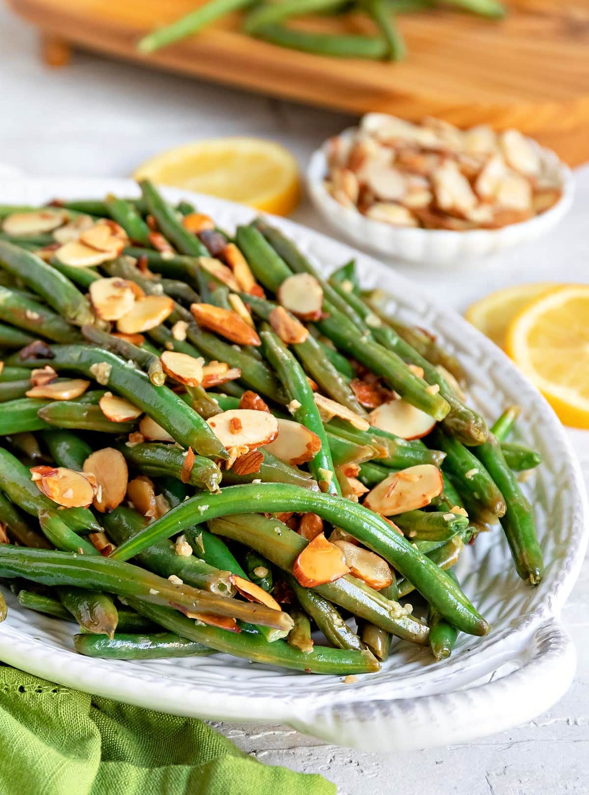 large white serving dish with green bean almondine ready to be served. topped with toasted almonds. small bowl of almonds in background next to two lemon slices.