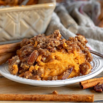 small plate with a serving of sweet potato casserole on it. Baking dish with the rest of the casserole in background. casserole is topped with a pecan streusel.