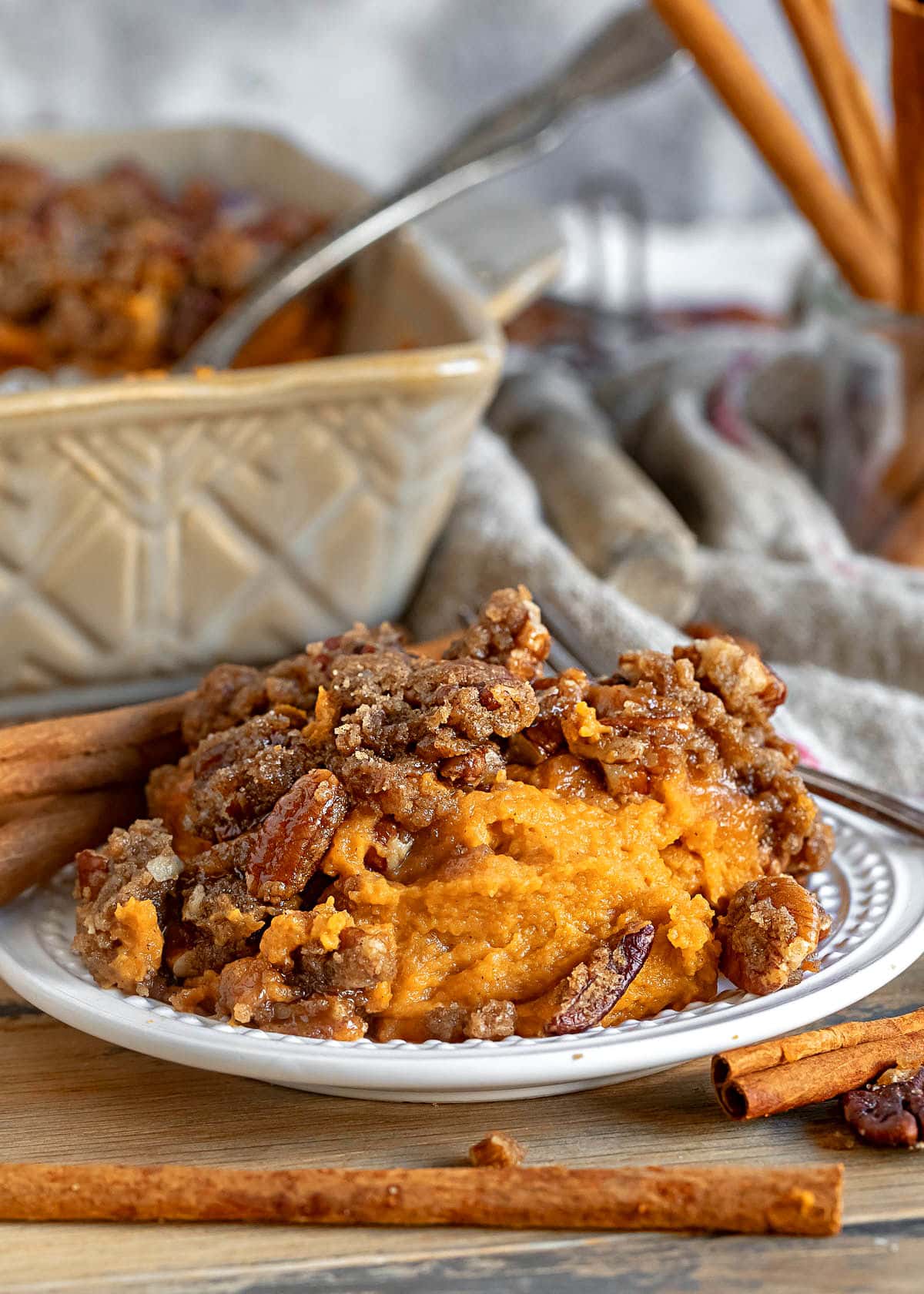 small plate with a serving of sweet potato casserole on it. Baking dish with the rest of the casserole in background. casserole is topped with a pecan streusel.