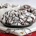 plate of chocolate crinkle cookies sitting on white wood board on red sparkly placemat. black and white twine sitting on board and more cookies can be seen in the background.