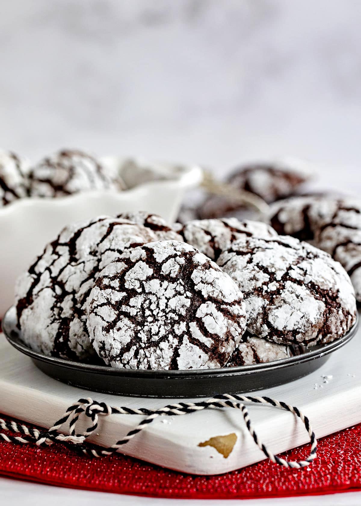 plate of chocolate crinkle cookies sitting on white wood board on red sparkly placemat. black and white twine sitting on board and more cookies can be seen in the background.