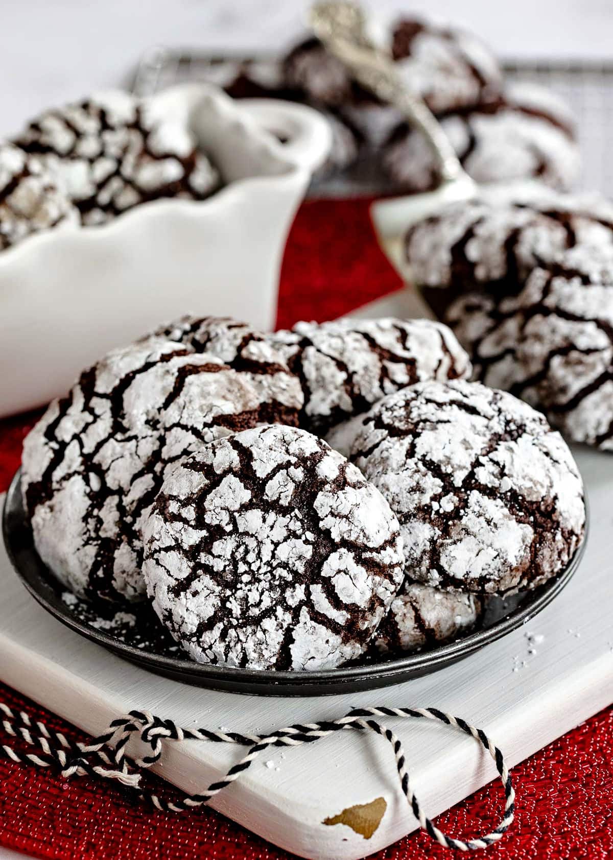 small metal plate with six chocolate crinkle cookies sitting on a white wood board with more cookies on a server and cooling rack in background.