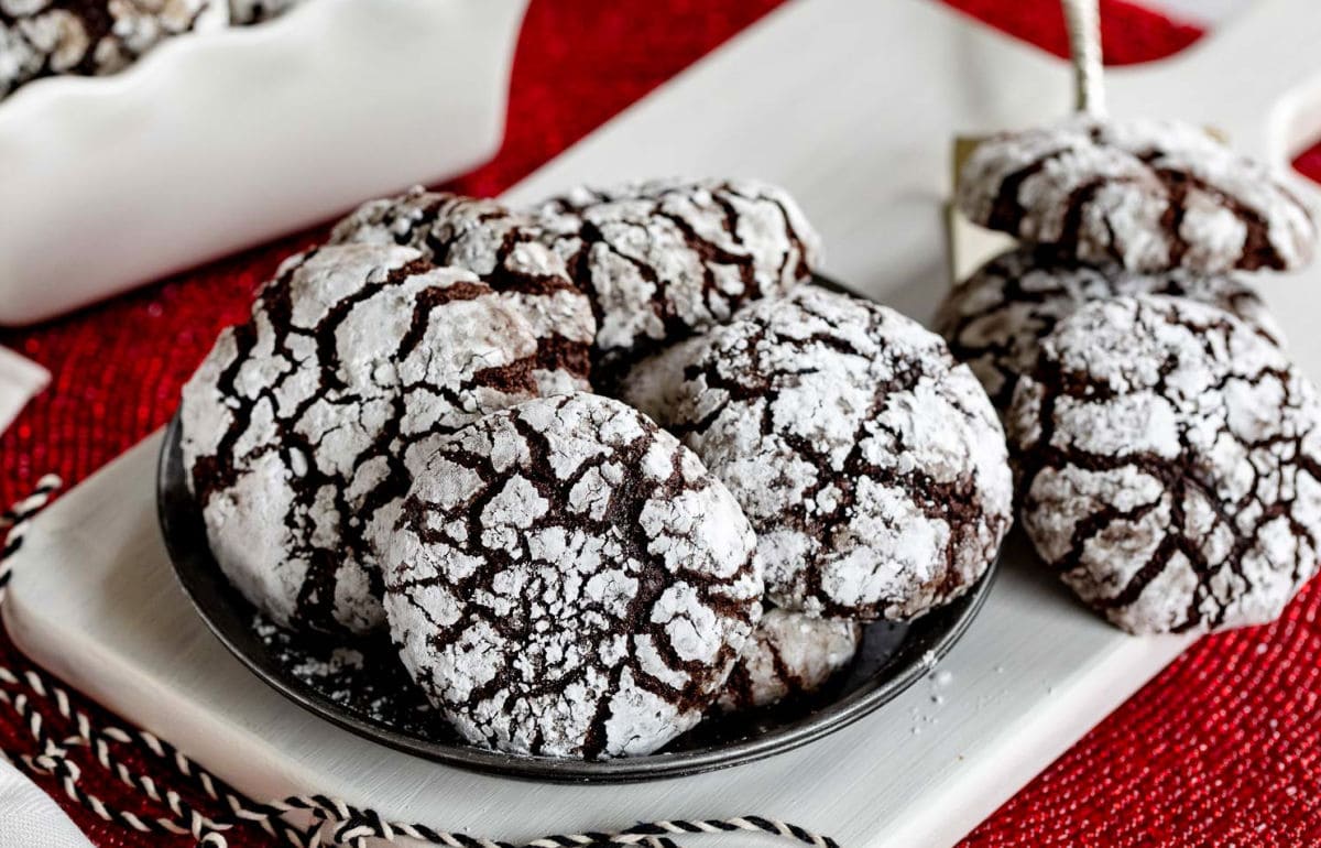 plate of chocolate crinkle cookies with more cookies sitting off to the side and a few more in a white ceramic container. red placemat sitting underneath.