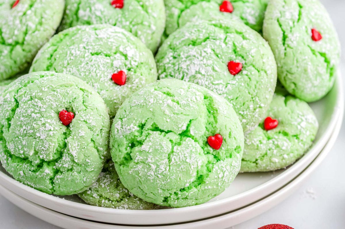 two white plates stacked with a pile of green grinch cookies with red heart sprinkles on top.