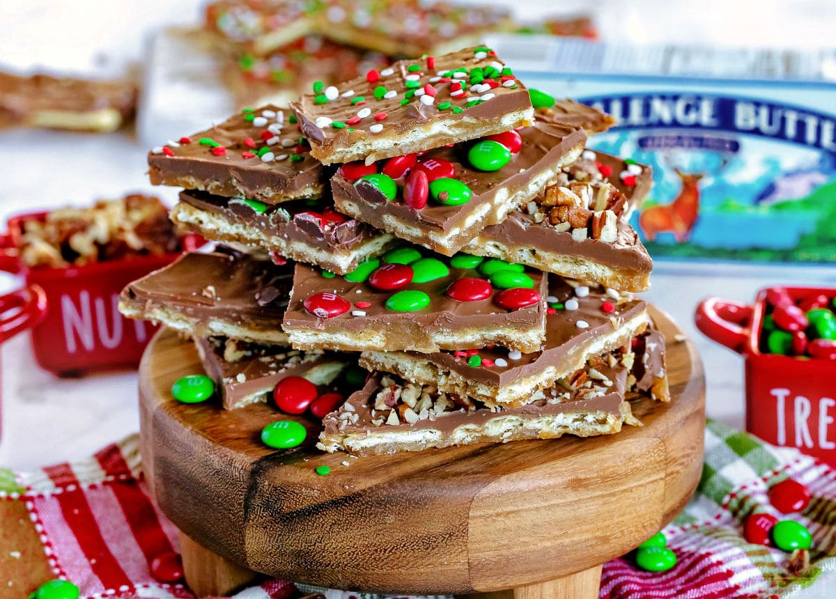 pieces of christmas crack saltine cracker toffee sitting on round wood board that is on a christmas napkin. toffee is decorated with sprinkles and m&ms.