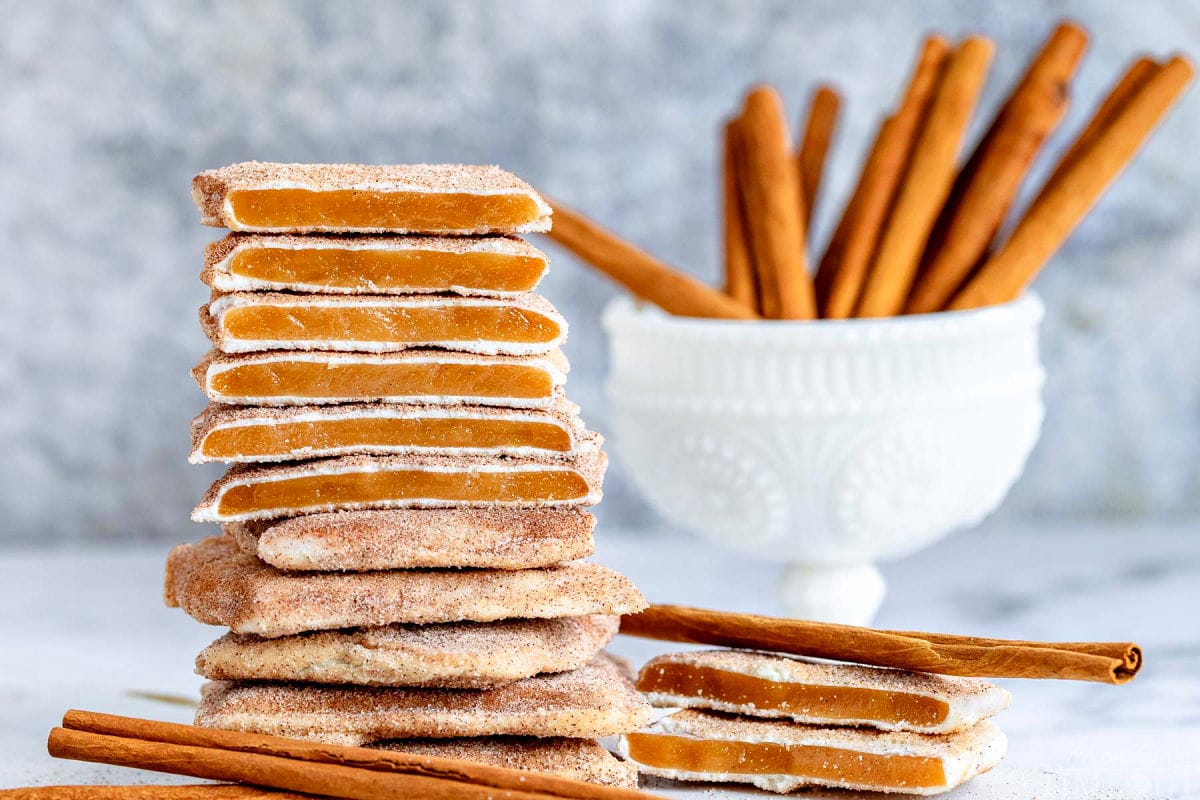 square churro toffee stacked on one another with several pieces broken in half so you see the toffee, the white chocolate layer and cinnamon sugar coating. cinnamon sticks in the background.
