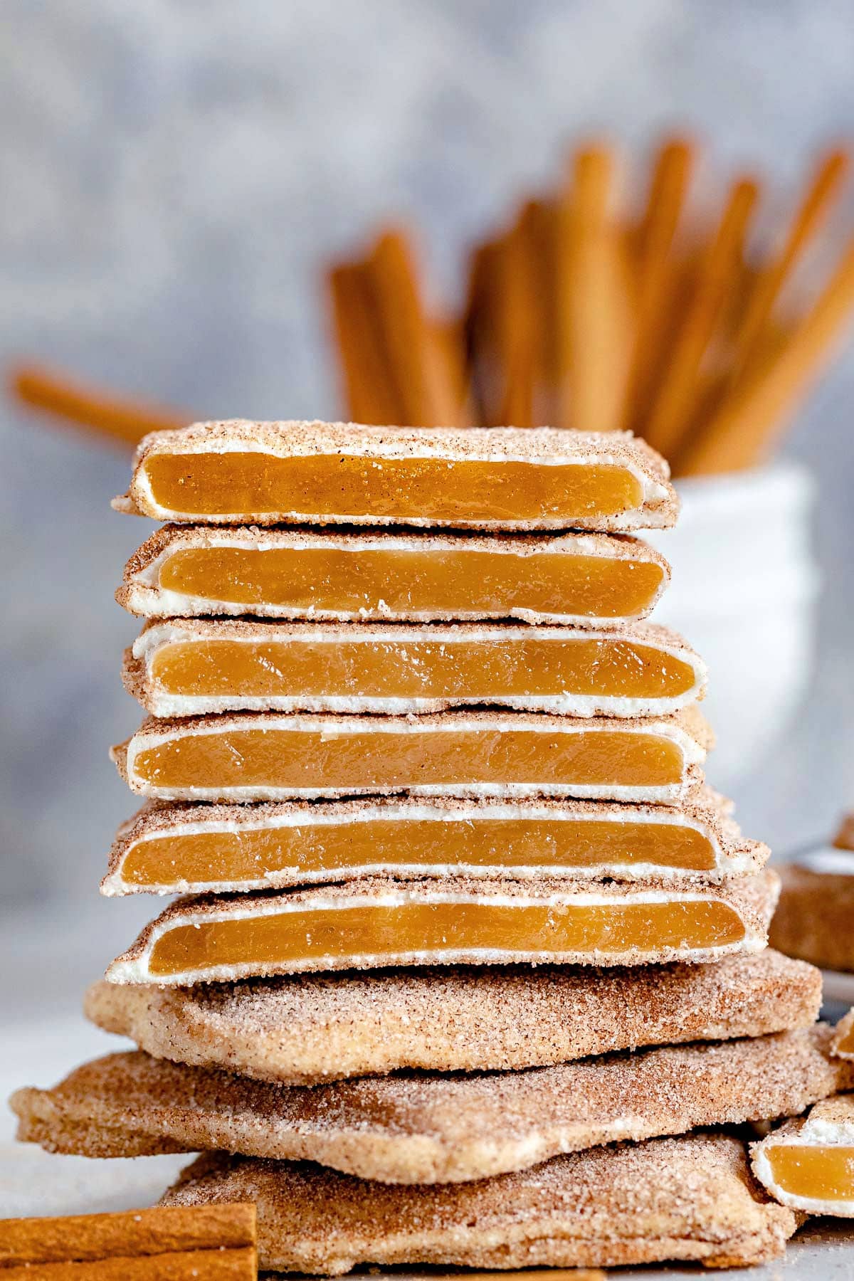 square churro toffee stacked on one another with several pieces broken in half so you see the toffee, the white chocolate layer and cinnamon sugar coating. cinnamon sticks in the background.