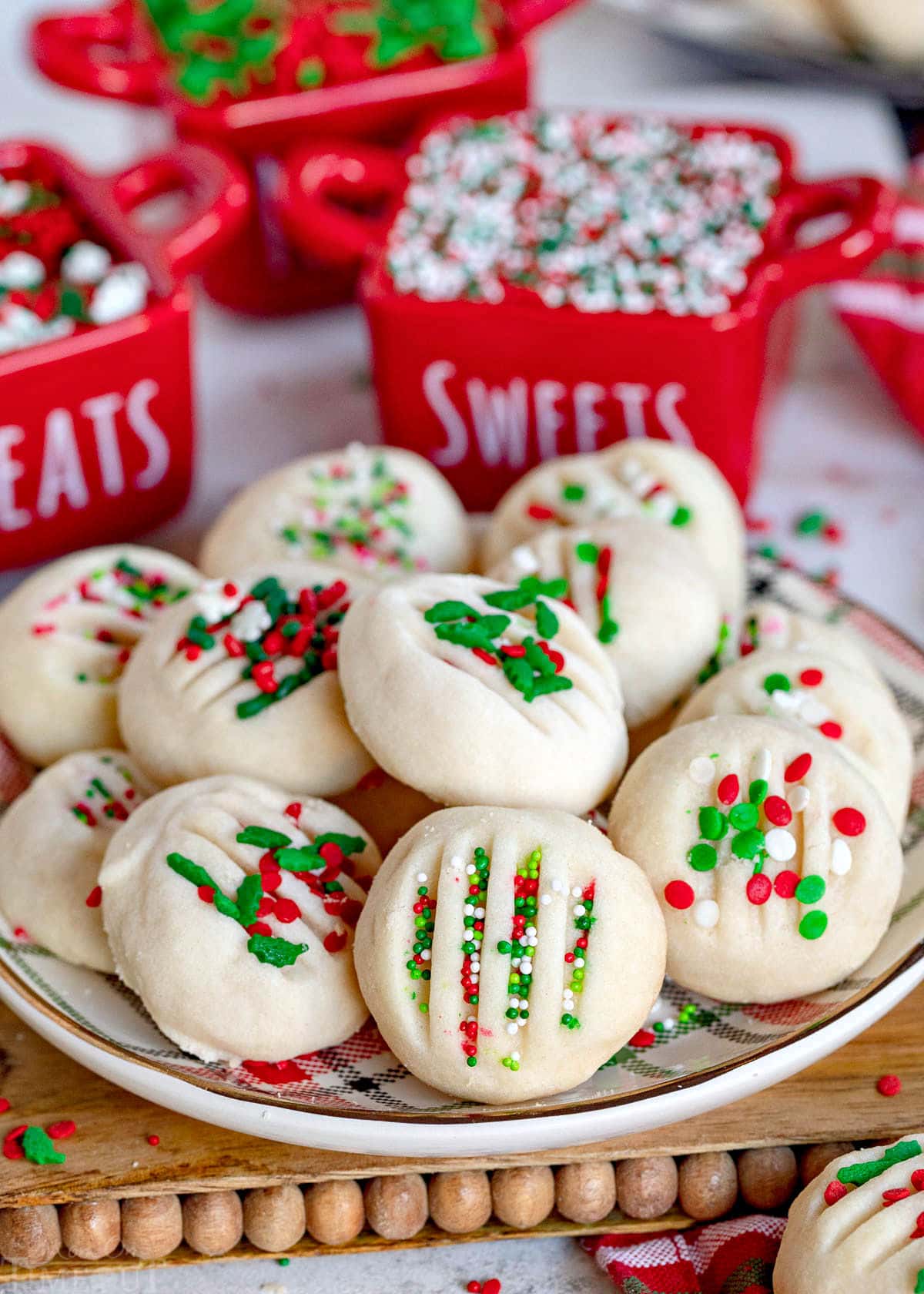 top down view of shortbread cookies on white plate decorated with lots of Christmas sprinkles. Small red jars are seen in the background filled with more sprinkles.