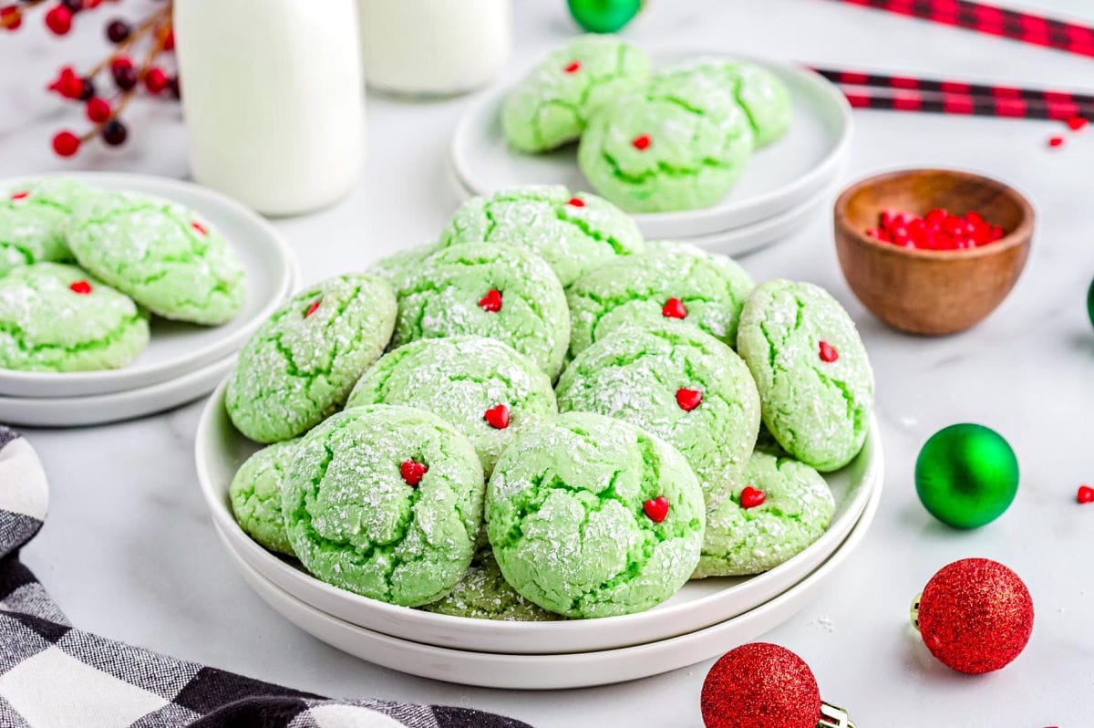 grinch cake mix cookies with red heart sprinkles sitting on white plate with milk jars in the background and Christmas ornaments scattered about.