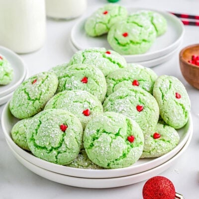 grinch cake mix cookies with red heart sprinkles sitting on white plate with milk jars in the background and Christmas ornaments scattered about.