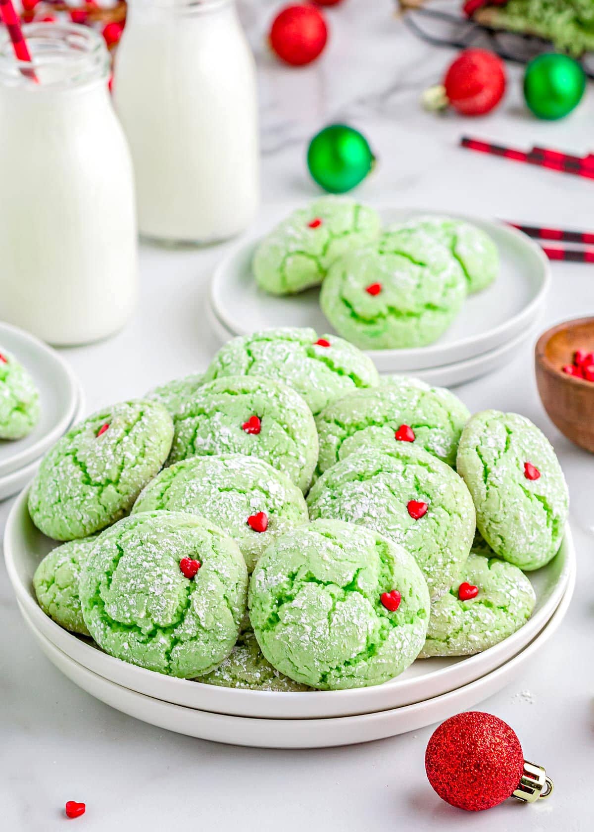 green cake mix cookies with red heart sprinkles sitting on white plate with milk jars in the background and christmas ornaments scattered about.