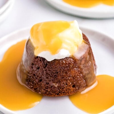 angled look at one sticky toffee pudding topped with whipped cream and more toffee sauce on a single white dessert plate.