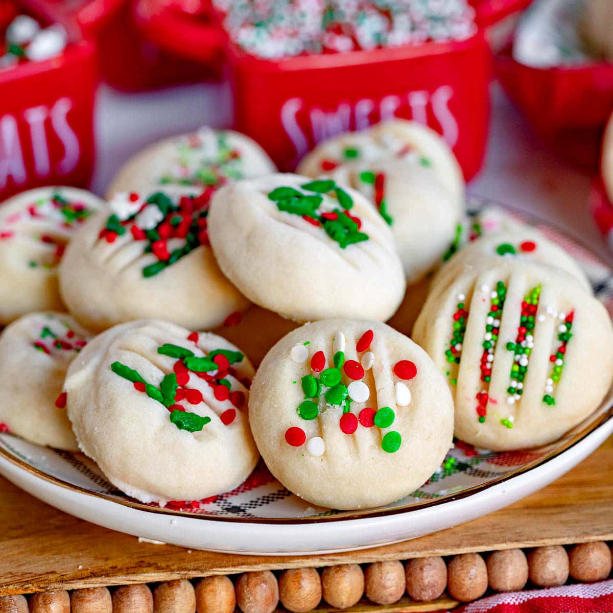 whipped shortbread cookies on a Christmas plate with red, green and white sprinkles. Plate is on cutting board sitting next to a Christmas towel.