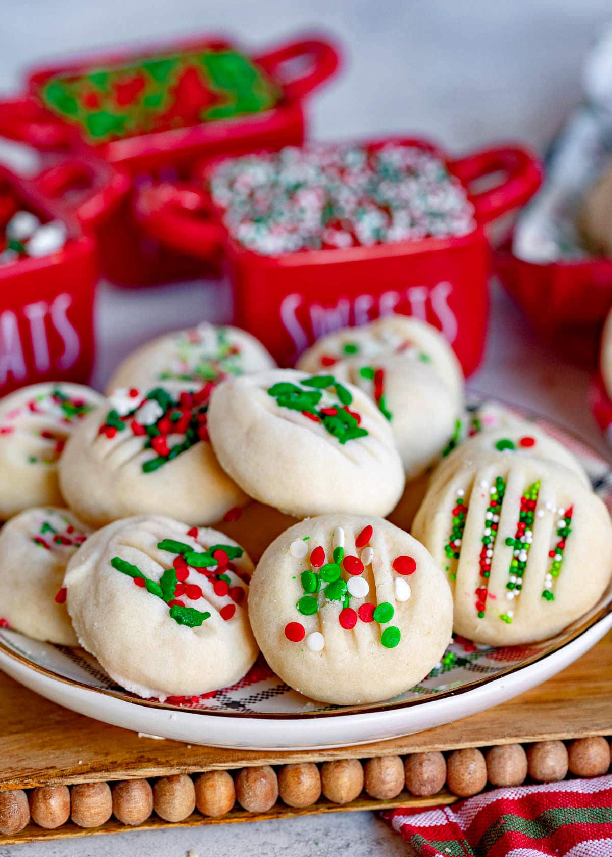 whipped shortbread cookies on a Christmas plate with red, green and white sprinkles. Plate is on cutting board sitting next to a Christmas towel.