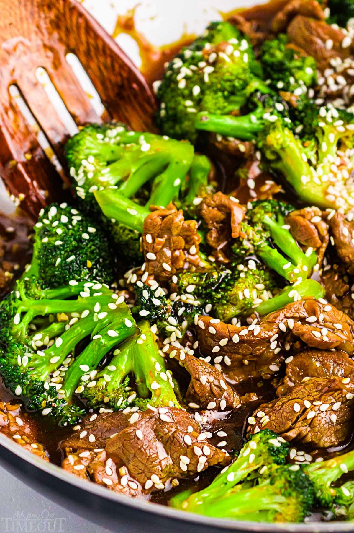 beef and broccoli stir fry topped with sesame seeds in white skillet with wood serving spoon.