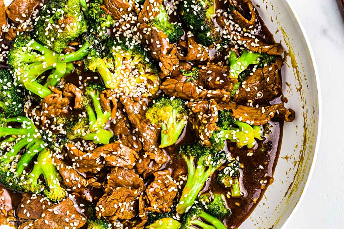 beef and broccoli stir fry recipe in white skillet topped with white sesame seeds. top down view.