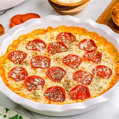 pizza dip baked in white pie dish topped with pepperoni and parmesan cheese. plate of toasted baguette and a bowl of chips are set off to the side.