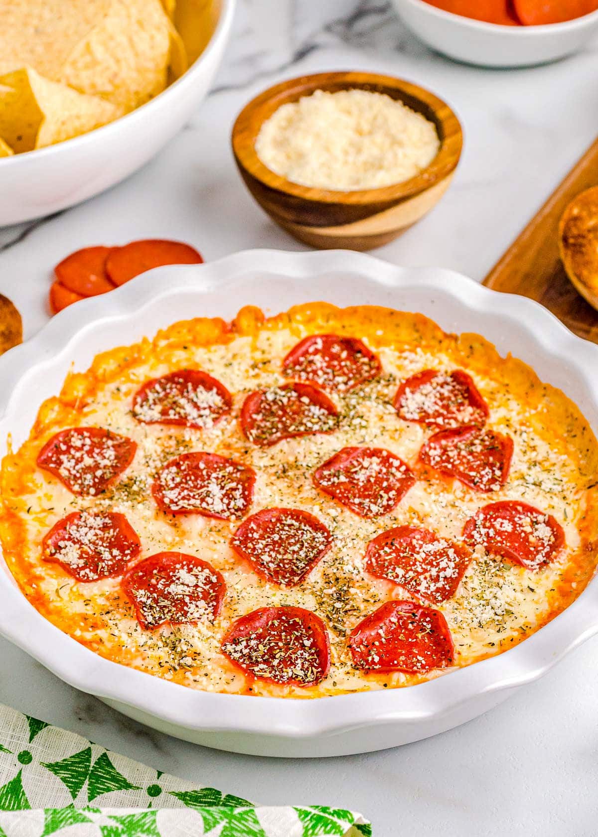 pizza dip baked in white pie dish topped with pepperoni and parmesan cheese. plate of toasted baguette and a bowl of chips are set off to the side.