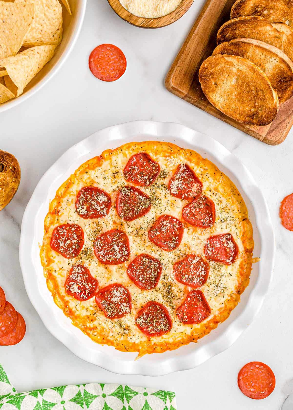 pizza dip baked in white pie dish topped with pepperoni and parmesan cheese. plate of toasted baguette and a bowl of chips are set off to the side.